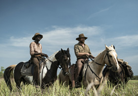 Two men on horses in a grassy field - Jacob Junior Nayinggul and Simon Baker in 'High Ground'. 